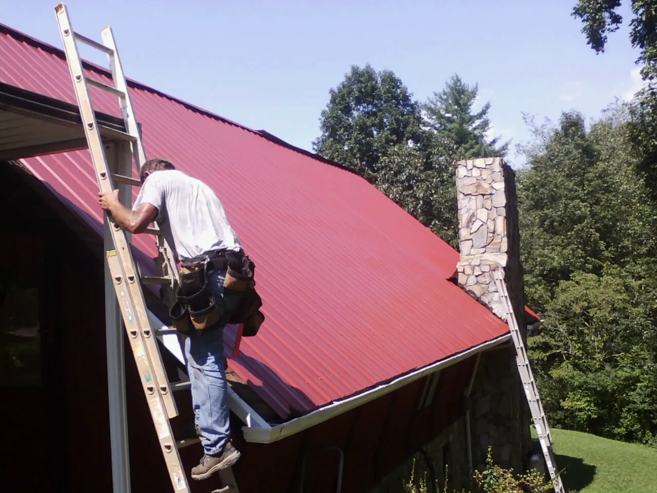 Roofer on a ladder beside a red metal roof and stone chimney — Soffit & Fascia Repair by Roofing Brothers in Arlington Heights